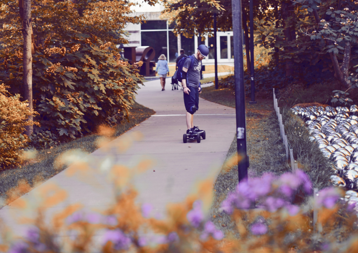A man riding a skateboard down a sidewalk, outside of the Crystal Bridges Museum of American Art in Arizona.
